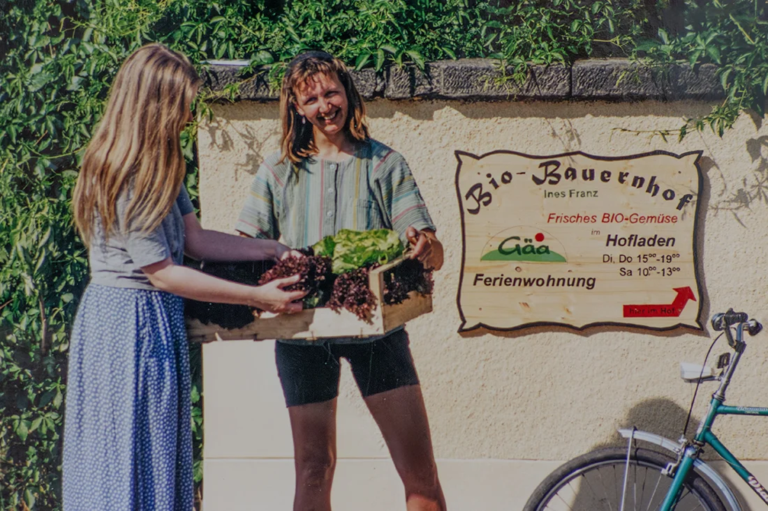 Ines Franz und eine junge Frau stehen vor dem Hofschild des Bio-Bauernhof Ines Franz; sie hält eine Kiste mit Salat